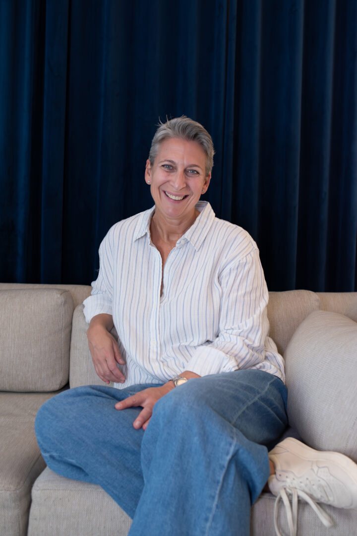Andrea Gartenmaier, Erfahrene Ordinationsplanerin in Wien, sitzend auf beigen Sofa, mit dunkelblauem Vorhang im Hintergrund