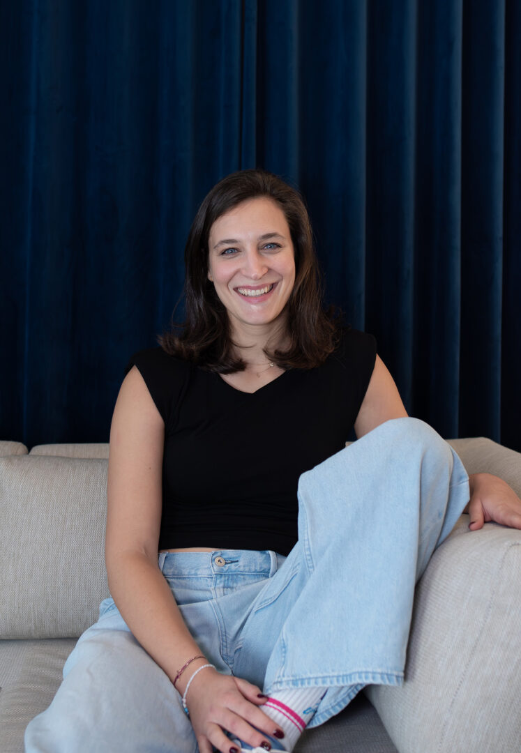 Team von fineconnect- Nicole Gartenmaier, Controlling und Key Account, sitzend auf beigem Sofa, mit dunkelblauem Vorhang im Hintergrund im Büro von fineconnect, Wien.